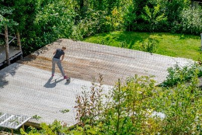 En kvinna behandlar trädäcket med träolja. Hon står med en trätvättare och en hink på ett delvis behandlat trädäck, omgiven av frodig grönska.